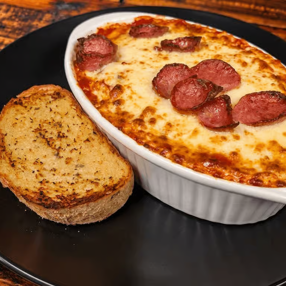 Baked cheesy pasta topped with sliced sausage, served with toasted garlic bread on a black plate atop a rustic wooden table.